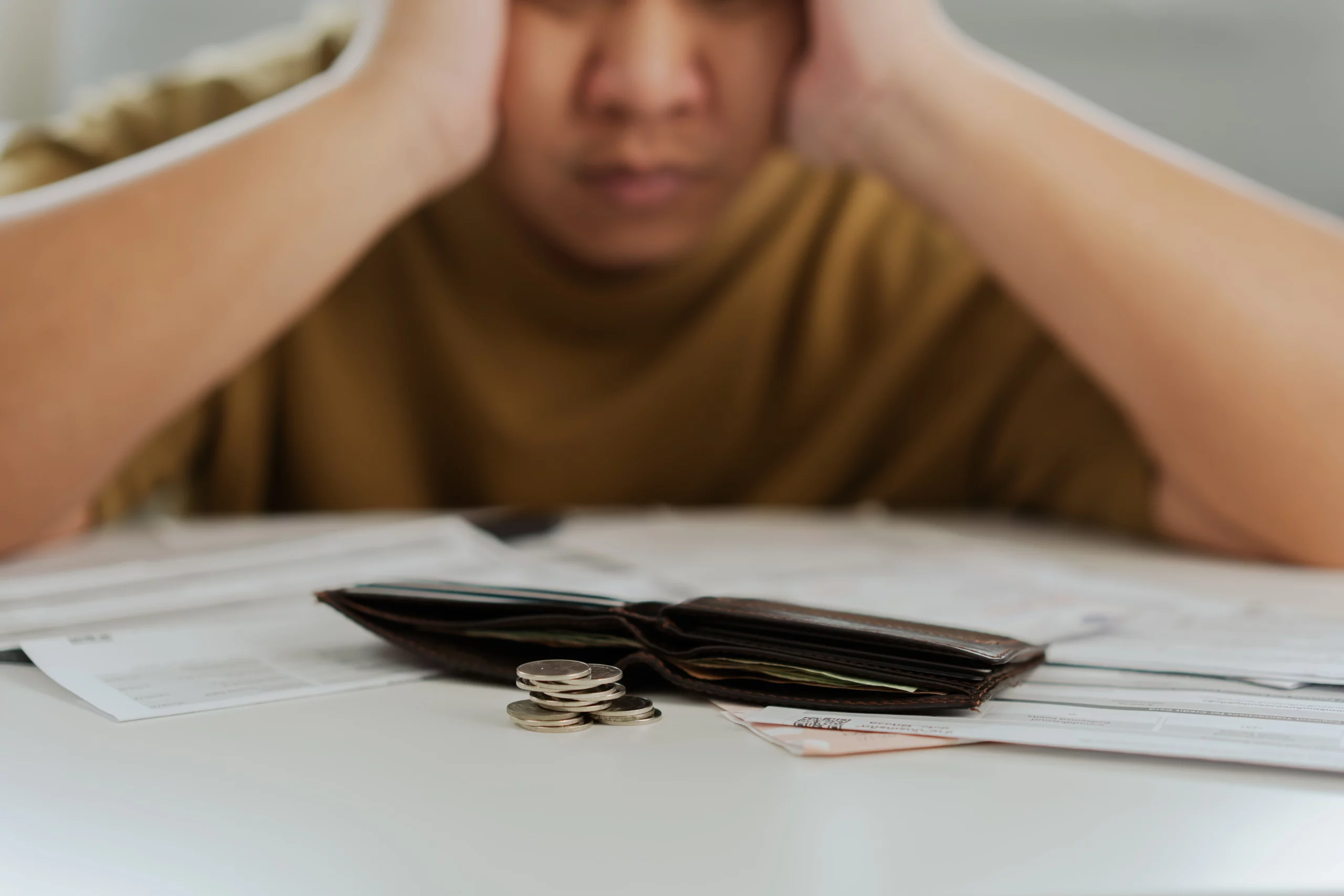 A worried person with their head in hands, surrounded by bills, an open wallet, and a small stack of coins, symbolizing financial stress.