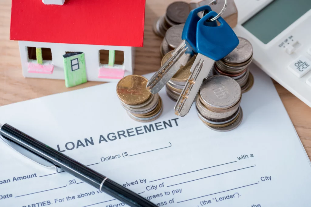 Miniature house with red roof beside stacks of coins, keys and loan documents on a wood table, symbolizing home financing and mortgage options.