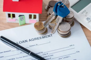 Miniature house with red roof beside stacks of coins, keys and loan documents on a wood table, symbolizing home financing and mortgage options.