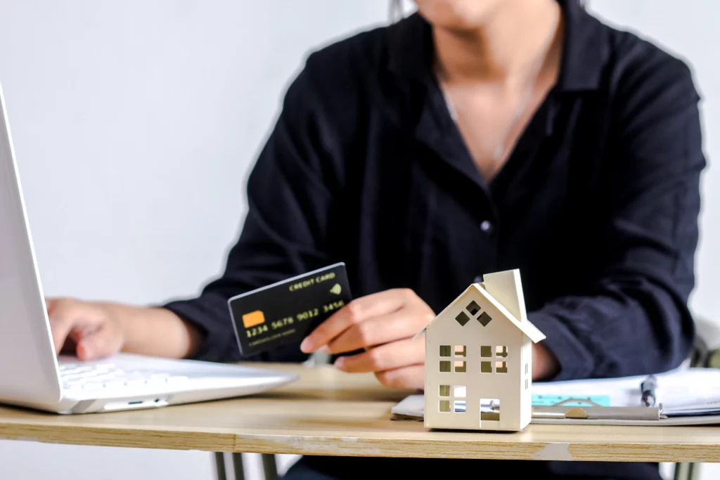 Hands using a credit card and laptop beside a wooden house model, illustrating budgeting and payment planning for a Maryland Chapter 13 bankruptcy repayment plan.